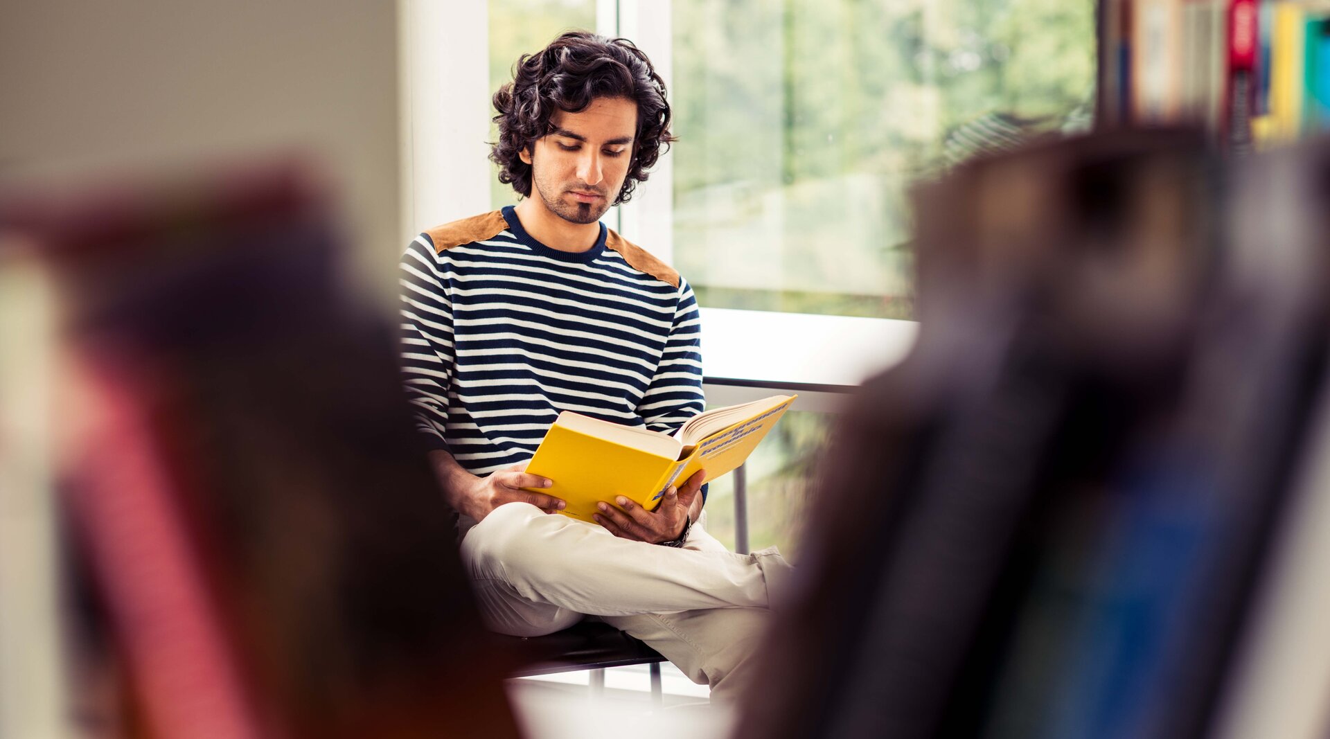Foto: Ein junger Mann sitzt auf einem Stuhl und liest in einem gelben Buch. Die Kamera blickt durch ein Bücherregal auf ihn.