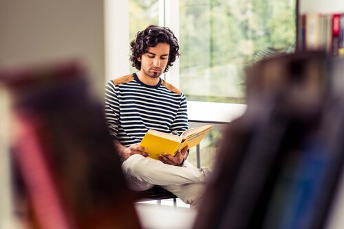 Foto: Ein junger Mann sitzt auf einem Stuhl und liest in einem gelben Buch. Die Kamera blickt durch ein Bücherregal auf ihn.